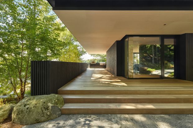 A modern black-clad building set among mature trees, viewed from a broad timber deck and stepped entrance. The deep roof overhang, dark vertical cladding, expansive glazed doorway, warm timber decking, and natural boulders create a refined connection to the surrounding woodland.