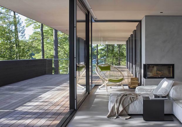 A refined modern living room opening onto a large timber deck through expansive floor-to-ceiling glazed doors. The room features a minimalist white sectional sofa, low dark side table, fireplace set into a smooth grey wall, and a pair of woven lounge chairs with green cushions. The deck is enclosed by dark privacy screening and overlooks a leafy wooded setting, creating a strong indoor-outdoor connection.