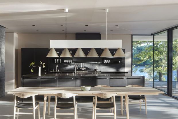 A spacious, refined open-plan kitchen and dining area. A long dark kitchen with a glossy black backsplash, integrated cabinetry, open shelving and a large island or peninsula occupies the rear wall. Multiple sculptural pendant lights hang above a long light timber dining table surrounded by contemporary dining chairs. Full-height corner glazing brings in abundant daylight and views of greenery, while a pale smooth ceiling and neutral walls create a clean, sophisticated setting.