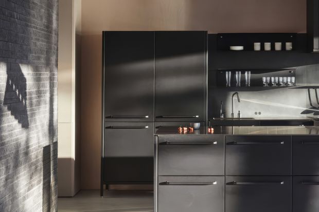 A compact, contemporary kitchen with dark matte cabinetry, a central island, integrated appliances, open shelving, and a stainless-steel sink and tap. A reflective splashback and glassware shelving sit beneath warm neutral walls, while strong sunlight casts shadows across a textured dark brick feature wall.