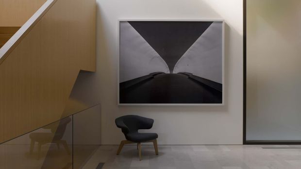 A minimalist interior corner beneath a staircase, featuring warm vertical timber wall panelling, a glass balustrade, pale smooth flooring, a sculptural black lounge chair, and a large monochrome framed artwork. A roller blind or dark-framed opening is visible on the right.