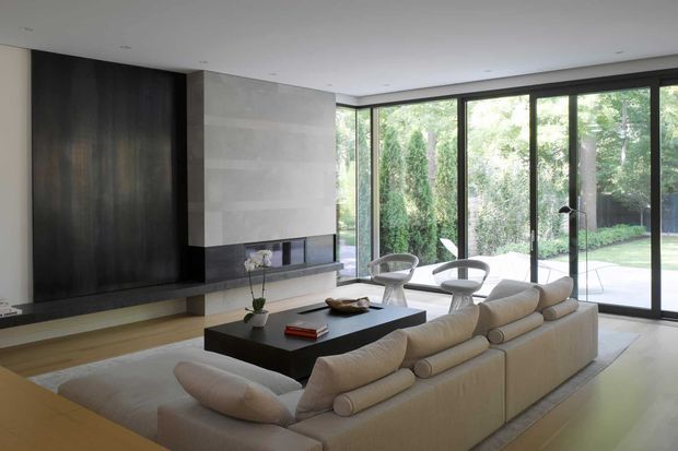 A refined contemporary living room with a large cream sectional sofa arranged around a low black coffee table and a minimalist built-in fireplace or media wall. Dark timber wall panelling contrasts with a pale stone or plaster feature surface, while expansive floor-to-ceiling glazing opens to a leafy garden. The room has a low, clean-lined ceiling, warm timber flooring, and a calm, neutral palette.