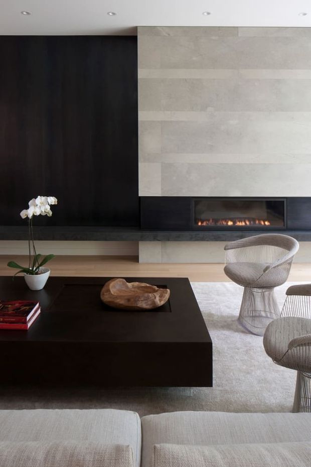 A sophisticated contemporary living room centered on a large dark coffee table, pale upholstered seating, sculptural woven lounge chairs, and a linear fireplace set into a dramatic black-and-light stone feature wall. Dark timber or black built-in cabinetry, recessed ceiling lights, a pale rug, and restrained decorative elements create a refined, high-contrast interior.