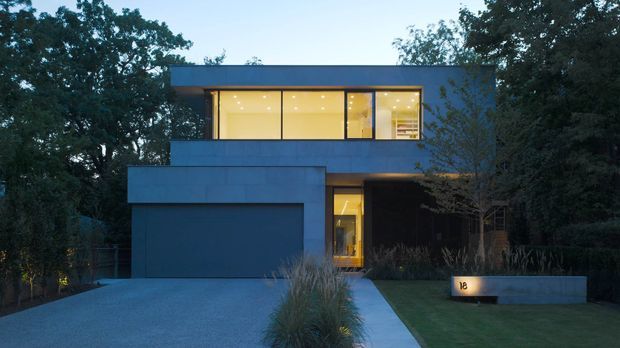 A dusk exterior view of a contemporary two-storey residence set among mature trees. The house has a simple rectilinear form with dark blue-grey cladding, a flat roof, extensive upper-level glazing glowing with warm interior light, an integrated garage, and a recessed front entry approached by a concrete path and driveway. Low landscaping and a rendered boundary wall frame the frontage.
