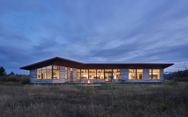 A wide dusk exterior photograph of a low, single-storey contemporary residence set in an open natural landscape. The home has a strongly horizontal composition, broad shallow overhanging roof, extensive warm-lit glazing, and a facade combining dark timber-like cladding with grey masonry or stone sections. A central glazed entrance and living spaces are visible from outside.