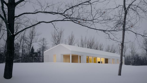 A minimalist white single-storey house set within a snowy, wooded landscape. The building has a simple low-pitched gable roof, clean white walls, and a long glazed facade with warm interior lighting visible through the windows and recessed openings. Bare deciduous trees frame the scene, creating a quiet winter setting.