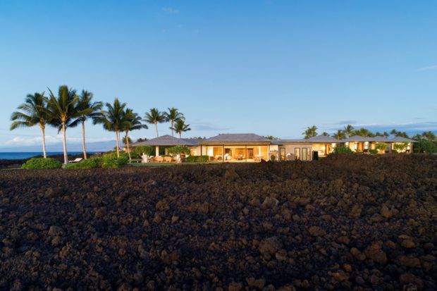 Wide exterior view of a low, sprawling single-storey residence set behind a broad field of dark volcanic rock. The home has a long, low-pitched roof, warm illuminated interiors, extensive glazing, and several tall palm trees around the property. Ocean and distant land are visible beyond the house under a clear blue sky, creating a tropical coastal setting.