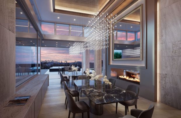Luxurious contemporary dining area with a long dark marble dining table, upholstered dining chairs, a linear built-in fireplace, and a dramatic suspended cluster chandelier. Floor-to-ceiling glazing opens to expansive sunset city views, while pale stone walls, warm timber ceiling detailing, and integrated lighting create a refined, elegant atmosphere. A large mirror reflects the dining space and chandelier.