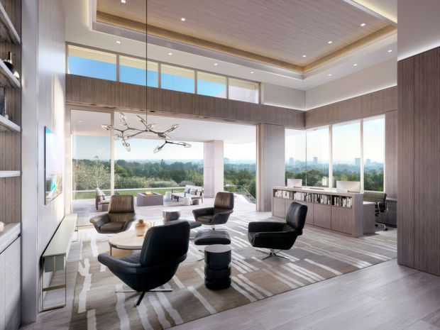 A bright, spacious contemporary living room with double-height glazing and expansive views over a wooded landscape. The room features a sculptural branching pendant light, several dark leather lounge chairs, a low round coffee table, a large patterned rug, built-in shelving, and a long dark timber-toned partition or media joinery. A compact workspace is visible at the right, while the ceiling combines recessed lighting with a warm timber-lined inset.