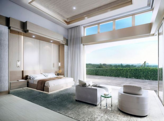 A spacious contemporary bedroom with a low-profile upholstered bed, integrated full-height timber-toned wall panelling and bedside joinery, and a pair of lounge chairs beside expansive floor-to-ceiling glazing. The room opens to a broad terrace overlooking a hedge and landscaped landscape. A recessed timber-lined ceiling feature with concealed detailing adds architectural interest. The palette is light and neutral with soft grey, white, and warm wood tones.