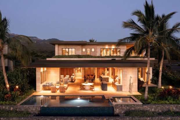 A luxurious two-storey modern tropical residence photographed at dusk, with warm interior lighting, broad glazed openings, timber-toned finishes, flat rooflines and an expansive covered outdoor living area opening onto a rectangular swimming pool and raised spa. Palm trees and landscaped planting frame the house, with rugged hills visible in the background.