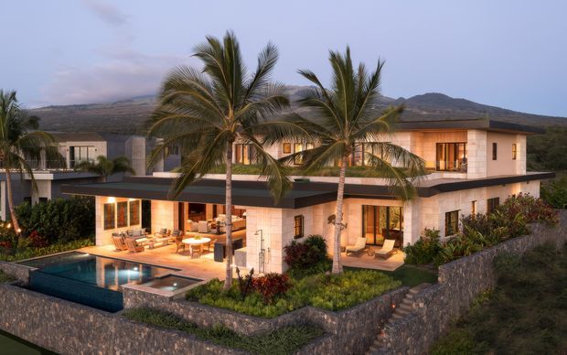 A large contemporary two-storey villa set in a tropical, mountainous landscape. The home has a low, flat roof, pale stone or rendered walls, extensive timber-framed glazing, warm interior lighting, landscaped gardens, a swimming pool and surrounding stone retaining walls.