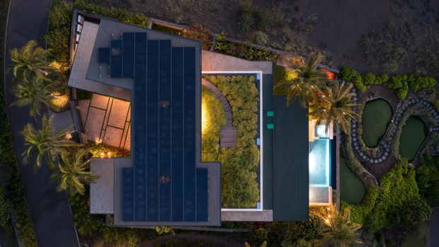 Aerial dusk view of a contemporary residential property with a large dark roof fitted with solar panels, landscaped courtyards, a swimming pool, palm trees, and winding ornamental ponds or garden water features.