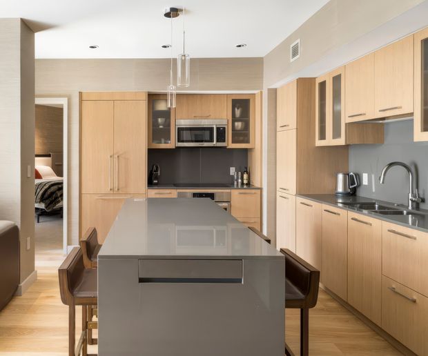 A contemporary galley-style kitchen with a large central island and integrated seating. Light timber cabinetry runs along the back and side walls, complemented by a dark grey splashback and polished grey island benchtop. Built-in appliances, under-cabinet storage, pendant lights, and a stainless-steel sink with gooseneck tap are visible. An open doorway at left reveals part of an adjoining bedroom.