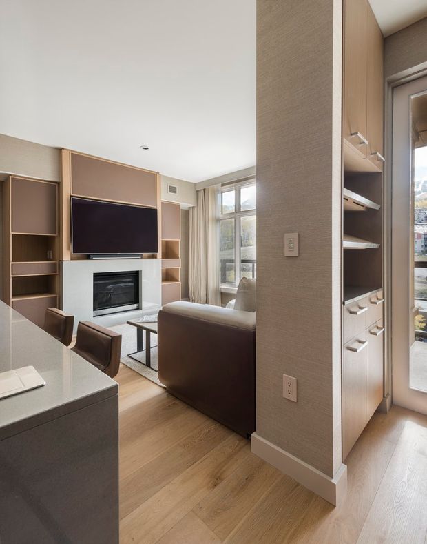 A warm, contemporary open-plan living area viewed from the kitchen. The room features a built-in timber-toned media wall with shelving, a wall-mounted television and fireplace, a brown sofa, and large windows with curtains. In the foreground is a pale stone kitchen island with dark cabinetry and dining stools. A tall built-in storage unit with open shelves and drawers lines the right side, and light timber flooring runs throughout.