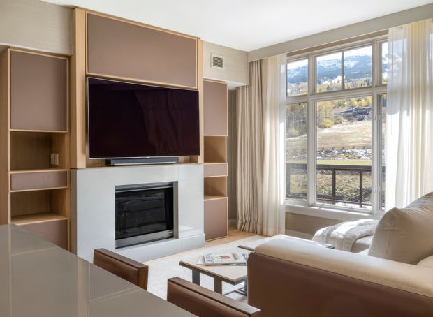 Bright contemporary living room with a built-in timber-toned entertainment wall, recessed television, electric fireplace, open shelving and pale cabinetry. A large multi-pane window with sheer curtains overlooks a wooded mountainous landscape, while a light upholstered sofa and low timber coffee table create a comfortable seating area.