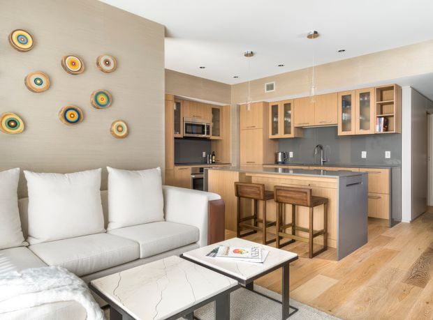Open-plan contemporary living and kitchen area with a light neutral palette, pale timber cabinetry and flooring, a dark grey kitchen island, integrated appliances, and a light upholstered sectional sofa. Colourful circular wall art adds small accents to the textured beige feature wall. The kitchen includes a peninsula with two timber stools, grey splashback or wall surfaces, pendant lights, and recessed ceiling lighting.