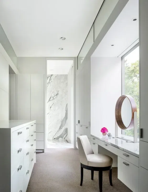 Bright, refined dressing room or walk-in wardrobe with extensive white built-in cabinetry and drawer storage on both sides. A long marble-topped vanity runs beneath a large round timber-framed mirror beside floor-to-ceiling glazing. The space has recessed ceiling lights, soft neutral carpet, and a dramatic white-and-grey marble wall visible through the opening at the rear.