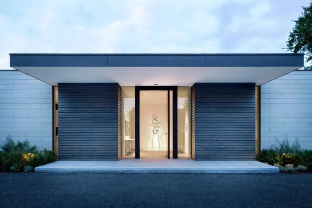 Front elevation of a low, single-storey contemporary residence with a broad flat roof and deep projecting eave. The symmetrical entry is framed by dark horizontal timber-like cladding panels and pale horizontal masonry or panel walls, with a glazed black-framed doorway opening into a softly lit interior. Minimal landscaping and a paved forecourt reinforce the clean modern composition.