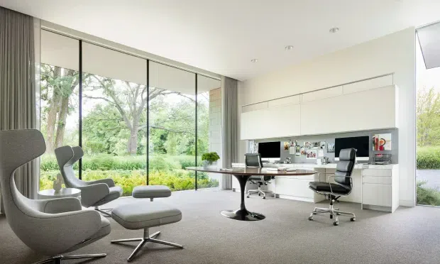 Bright, spacious contemporary home office with floor-to-ceiling glazing overlooking a leafy garden. The room includes a large built-in white workstation with integrated cabinetry, two office chairs, a central desk, and a sculptural lounge chair with matching ottoman. Neutral finishes and abundant natural light create a calm, professional setting.