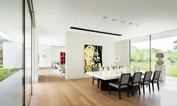 Bright, spacious contemporary dining area with a long white dining table and black upholstered chairs, set within an open-plan interior. Full-height glazing on two sides overlooks landscaped grounds, while pale timber flooring, white walls, recessed downlights, and a sculptural artwork create a refined modern atmosphere. A living area is visible beyond the central partition.