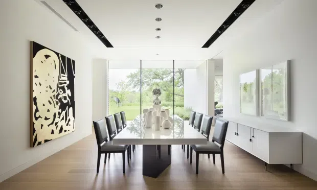 A bright, minimalist dining room with a long rectangular dining table surrounded by black dining chairs. The room has pale timber flooring, white walls, a streamlined white sideboard, abstract monochrome wall art, recessed ceiling lighting, and expansive floor-to-ceiling glazing overlooking greenery.