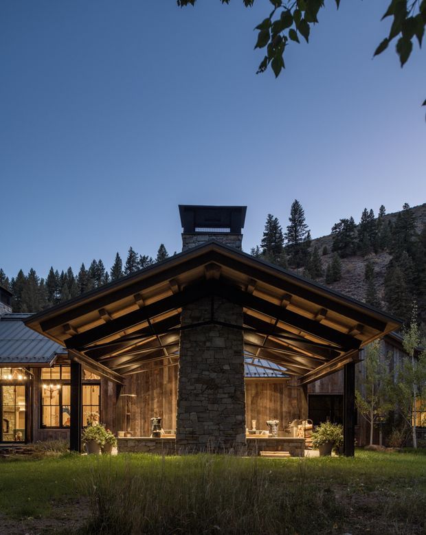 Exterior view of a rustic mountain residence at dusk, centered on a large open-sided gabled structure with exposed timber rafters, warm integrated lighting, and a substantial rough-stone chimney or fireplace column. Glazed and timber-clad living spaces sit to either side, with conifer-covered hills behind the property.