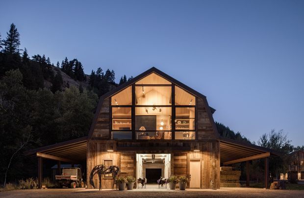 A warmly illuminated two-storey barn-style building at dusk, set against a wooded hillside. The timber-clad facade features a prominent glazed gable revealing an upper-level interior, a central entrance, broad low-pitched side roof extensions, and rustic outdoor landscaping.