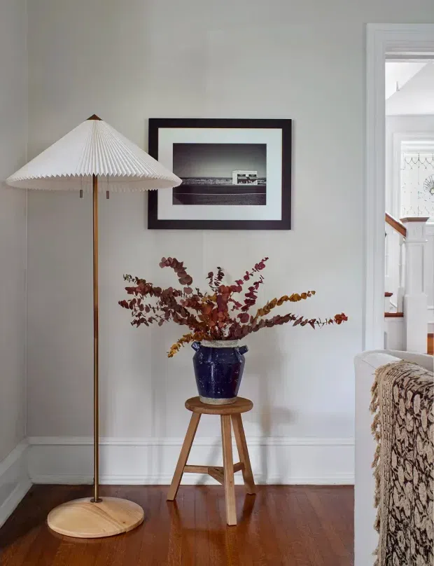 A styled residential interior corner with white walls and trim, polished dark timber flooring, and a tall pleated-shade floor lamp. A blue ceramic vase filled with dried branches sits on a small wooden stool beneath framed artwork. At right, a doorway opens toward a stair hall with white balustrades and timber handrails, while the edge of a patterned upholstered sofa is visible in the foreground.