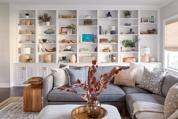 Bright, refined living room with a large grey sectional sofa positioned in front of extensive white built-in shelving and lower cabinetry. The shelves display books, ceramics, baskets, plants, and decorative objects, while layered cushions, a textured rug, timber side tables, and a central coffee table create a warm, inviting setting. A window with a light roller blind is visible on the right.