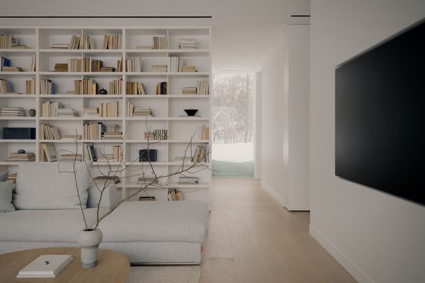 A bright, minimalist living room with a large white built-in bookshelf covering the rear wall, filled with books and restrained decorative objects. A pale modular sofa occupies the foreground beside a low rounded wood coffee table, while a wall-mounted television is visible on the right. Light timber flooring, white walls, concealed linear ceiling lighting, and a glazed opening toward a softly landscaped exterior create an airy, contemporary setting.