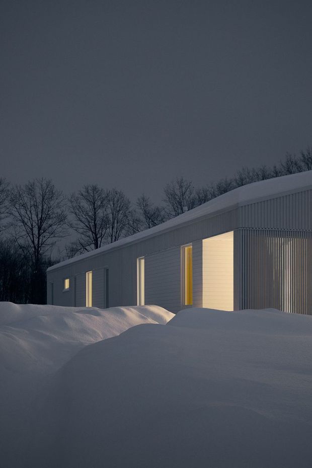 A minimalist single-storey building viewed externally at dusk, partially surrounded by deep snowdrifts. The long rectangular structure has a gently sloping roof, dark grey vertically ribbed cladding, narrow illuminated windows and doors, and a large warm-lit glazed or open bay screened by vertical fins. Leafless trees form a subdued background, creating a quiet winter architectural scene.