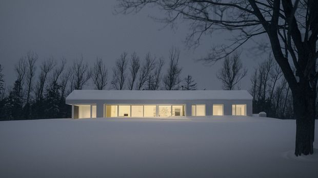 A low, elongated modern house viewed at dusk through a broad snow-covered foreground. The building has a simple flat or very low-pitched roof, a restrained pale facade, and a continuous band of warmly illuminated windows and glazed doors. Bare deciduous trees frame the scene, creating a quiet, secluded winter setting.