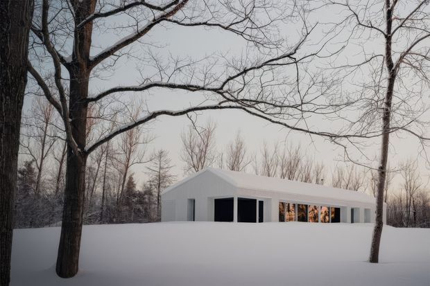 A minimalist single-storey white pavilion-like house set within a snow-covered wooded landscape. The building has a low, simple pitched roof, clean white walls, and extensive dark-framed glazing and openings. Bare deciduous trees frame the foreground and surround the structure, creating a quiet, stark winter scene.
