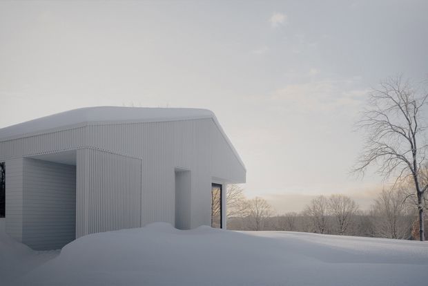 A minimalist white, single-storey building partially buried in deep snow, with corrugated vertical cladding, a gently curved roof edge, and dark-framed openings. The structure sits in a quiet wooded winter landscape with bare trees and soft overcast light.