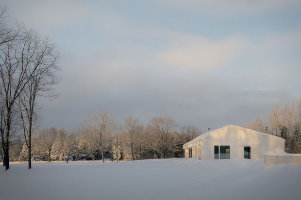 A minimalist white single-storey building sits in a broad snow-covered landscape, surrounded by leafless trees. The simple gabled roof, pale facade, dark rectangular windows, and soft overcast winter light create a quiet, secluded architectural exterior scene.