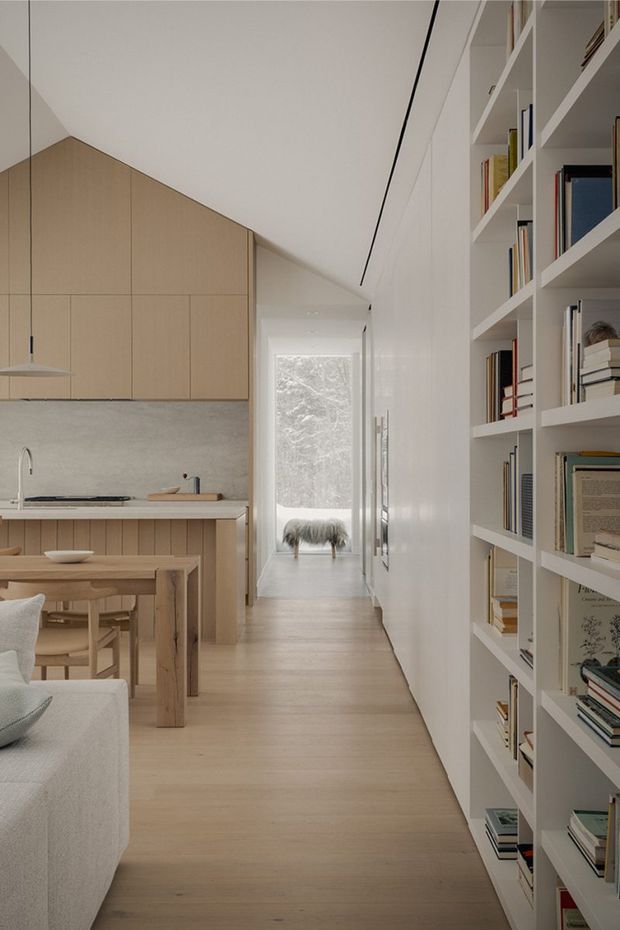 A bright, narrow open-plan interior combining a minimalist kitchen, dining area, living space and hallway. Pale timber cabinetry and flooring contrast with a light grey stone or concrete-look splashback, while a full-height white built-in bookshelf lines the right wall. Soft daylight enters through a glazed opening at the rear, creating a calm, warm and highly ordered atmosphere.