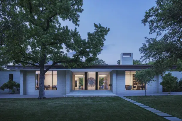 Single-storey modern residence viewed from the front across a lawn at dusk. The low, horizontal composition has pale masonry walls, extensive floor-to-ceiling glazing, a dark flat roof with a raised rectangular chimney or clerestory element, and a central recessed entry reached by a short set of steps. Mature trees frame the house and a simple stepping-stone path leads from the lawn to the entrance.