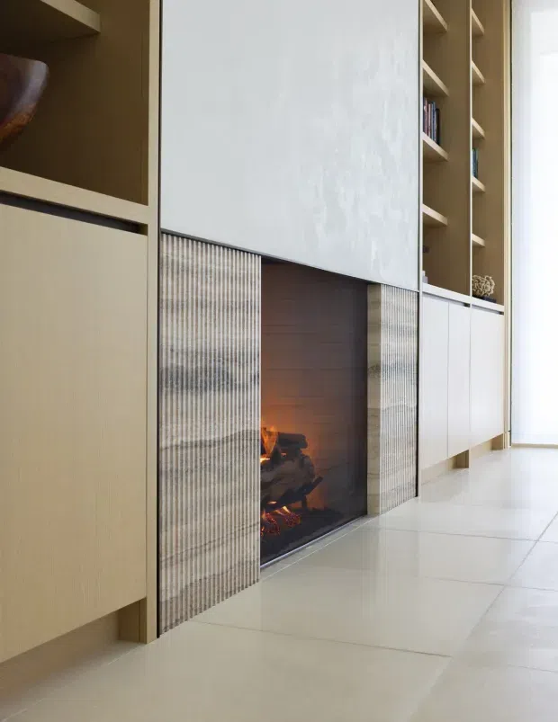 Built-in contemporary living-room joinery with a central fireplace, pale wood cabinetry, open shelving, white panel fronts, and vertically textured sliding screens. Large pale floor tiles and bright natural light create a clean, refined interior.