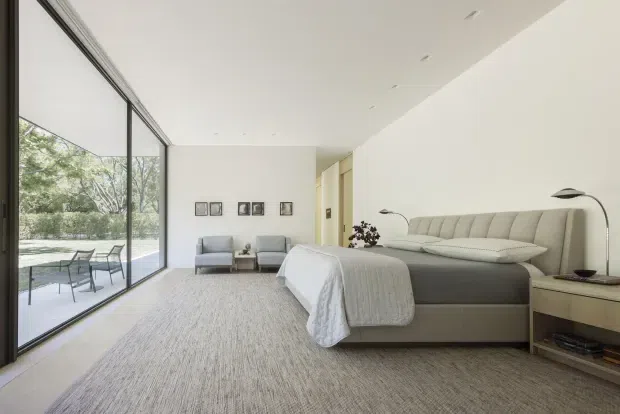 A bright, spacious contemporary bedroom with a large upholstered bed, neutral bedding, two armchairs, a small side table, bedside cabinetry, and slim floor lamps. Full-height glazing opens visually to an outdoor terrace and leafy landscape. The room has a restrained palette of white, cream, beige, and light grey, with a soft textured carpet and minimalist detailing.