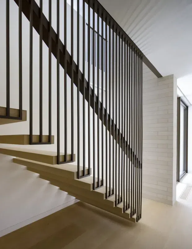 Interior view of a contemporary stairway with floating timber treads, slender dark metal vertical balusters, and a diagonal handrail forming a sculptural screen. The stair is set against white walls and a pale horizontal brick feature wall, with large glazing to the right and a light timber floor below.