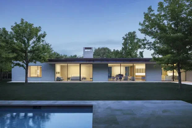 Single-storey modern residence viewed from the landscaped front or poolside exterior at dusk. The long, low rectangular form has a flat roof, dark roof fascia, extensive floor-to-ceiling glazing, and pale blue-grey masonry or rendered wall sections. Warm interior lighting reveals living and dining spaces behind the glazing. A reflecting pool and broad concrete-paved terrace occupy the foreground, with mature trees framing the property.