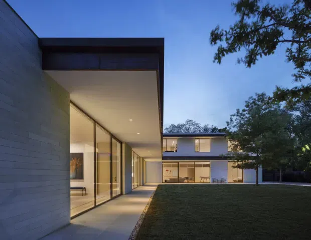 Twilight exterior view of a contemporary, flat-roofed two-storey residence organised around a broad lawn courtyard. The foreground wing has a deep projecting roof canopy, pale masonry walls, extensive floor-to-ceiling glazing, and a covered concrete walkway. A second-storey volume and illuminated ground-floor living spaces are visible beyond the lawn, with mature trees framing the property.
