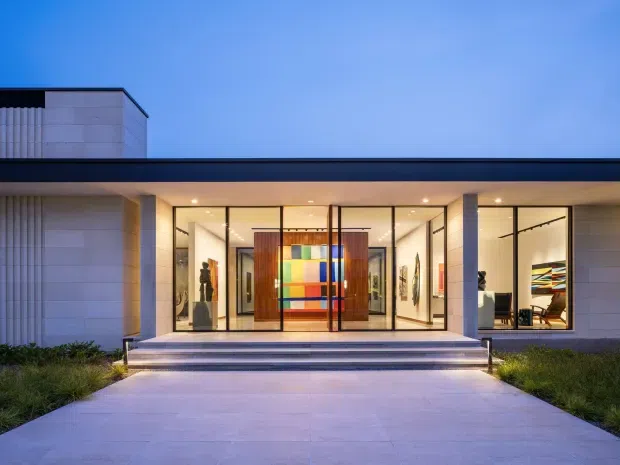 A contemporary flat-roofed building facade photographed at dusk, with a broad glass-fronted entrance and gallery-like interior visible through the glazing. Pale stone or concrete cladding frames the long dark roof edge, while wide steps lead from a paved forecourt to the illuminated entry. Artwork and furnishings are visible inside, including a colourful central feature panel.