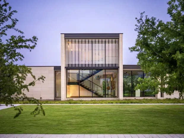 Front exterior view of a low-rise contemporary building with a symmetrical central glazed stairwell pavilion, pale stone or masonry wall panels, extensive floor-to-ceiling glazing, and a flat roofline. The building is set behind a broad manicured lawn and framed by mature trees, with warm interior lighting visible through the glass.