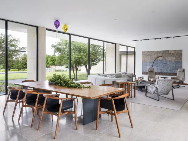 Open-plan contemporary dining and living area with a large timber dining table and mid-century-inspired wooden chairs in the foreground. Floor-to-ceiling black-framed glazing provides expansive views of the landscaped grounds, while a pale stone feature wall, light upholstered seating, and a large abstract artwork define the living zone. The room has a bright, refined, indoor-outdoor character with a high ceiling and strong natural light.