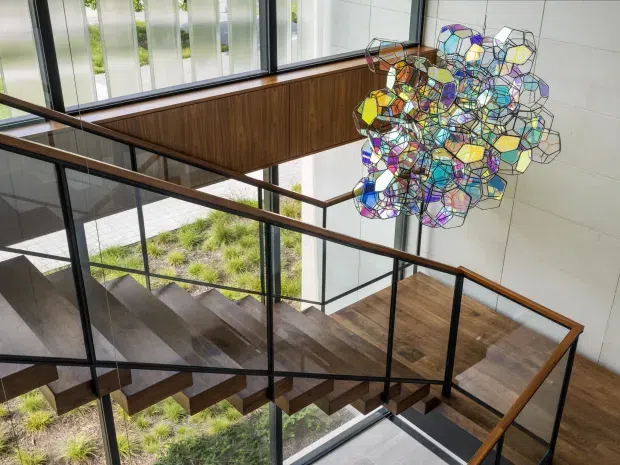 A modern stairway viewed from an upper landing, featuring dark timber stair treads and flooring, slender black metal and glass balustrades, warm timber wall panelling, and expansive floor-to-ceiling glazing. A large colourful geometric pendant light hangs in the stair void, with landscaped planting visible outside.