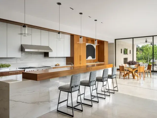 Bright open-plan contemporary kitchen with a long timber island and waterfall-style light stone ends, white cabinetry, marble-look splashback, stainless-steel rangehood and pendant lighting. A row of bar stools faces the island, while a glazed dining area with timber furniture and garden views sits beyond a black-framed glass partition. The space has a clean, refined, airy feel with warm timber accents.