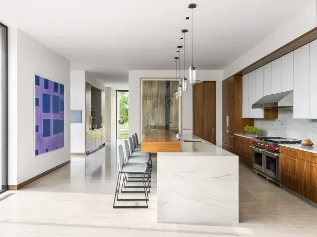 Bright, spacious contemporary kitchen with a large white stone island, integrated dining extension, and a row of slim upholstered stools. The kitchen features white flat-panel overhead cabinetry, warm timber lower cabinetry and tall storage, stainless-steel appliances, a pale tiled or stone floor, and a marble-look splashback. Linear pendant lights hang above the island, while a colourful geometric artwork and glazed internal partition add visual interest. An adjoining hallway and outdoor greenery are visible beyond the kitchen.
