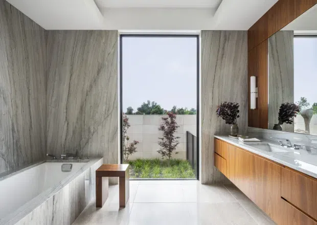 Bright contemporary bathroom with a freestanding white bathtub set against large grey-veined stone-look wall panels, a full-height glazed opening to a planted courtyard, and a timber vanity with integrated double basins beneath a large mirror. The space has a clean, refined material palette with white surfaces, grey stone tones, and warm wood cabinetry.
